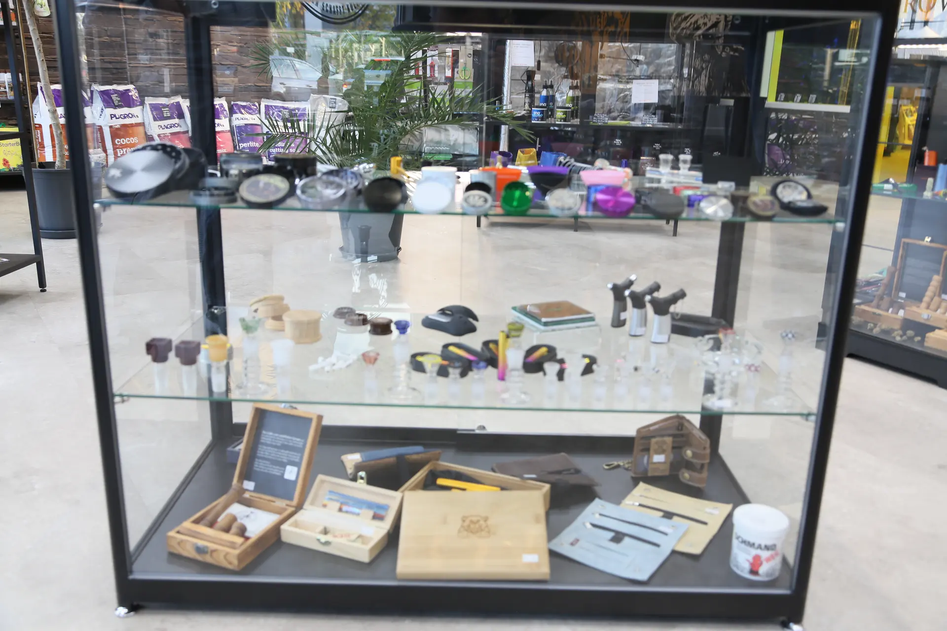 Glasvitrine mit drei Böden: oben Grinder, in der Mitte Glasköpfe und Feuerzeuge, unten Holzboxen und Taschen